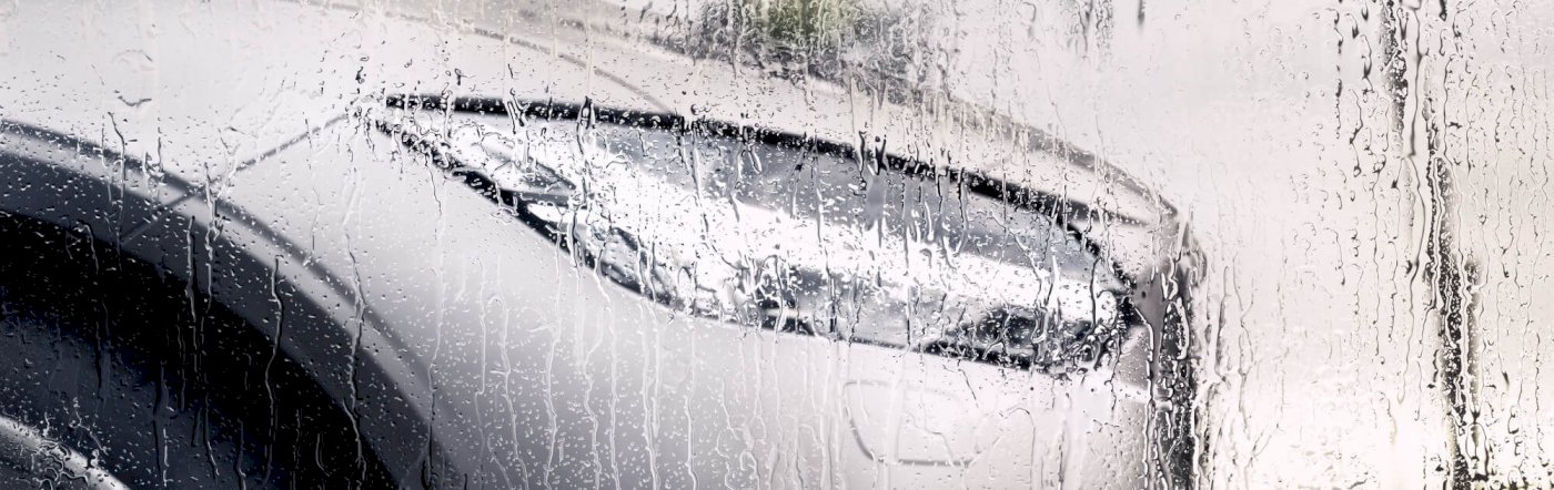 Die Schnauze des Volvo S90 INSCRIPTION in Bright Silver metallic steht vor einer nassen Scheibe, im Hintergrund ist ein Baum zu erkennen - Seitenschuss